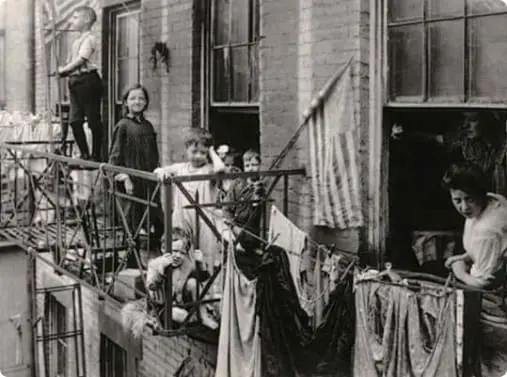 Black and white photo of historic tenement apartments with residents visible on narrow balconies. Laundry hangs on clotheslines strung across the building, capturing everyday life in a densely populated urban neighborhood.