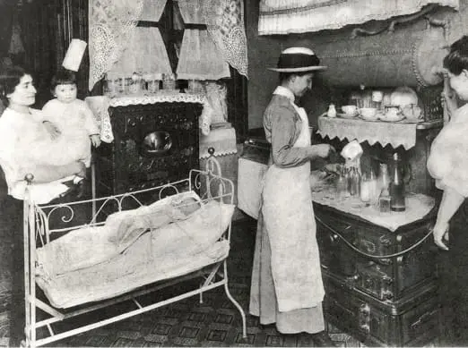 In a tenement kitchen, a VNS Health nurse educates a mother, holding her baby, how to prevent illness.