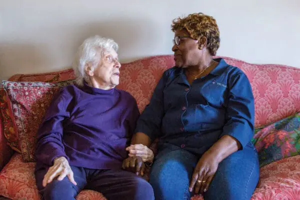A VNS Health home health aide sits next to her patient on a red sofa.