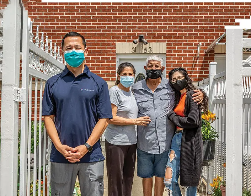 A VNS Health team member stands at a safe distance from a patient and their family. Everyone is wearing masks, reflecting safety measures during the COVID-19 pandemic.