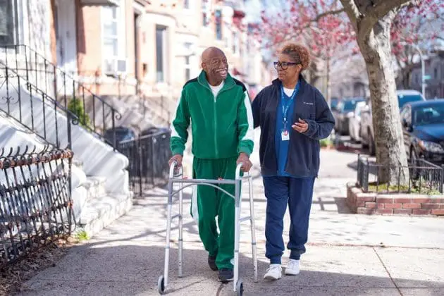 A VNS Health home health aide walks outside with a senior man who needs the support of a walker.