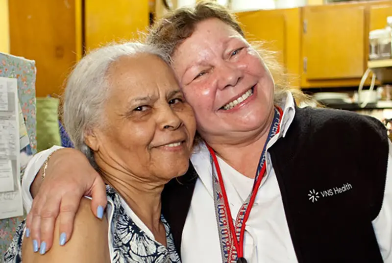 A VNS Health home health aide hugging her patient and smiling.
