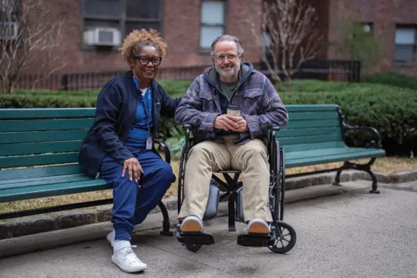 A VNS Health home health aide sitting next to her patient in a wheelchair.