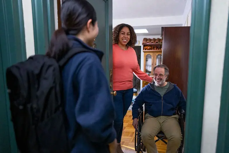 A VNS Health patient in a wheelchair and caregiver greet a team member at door.