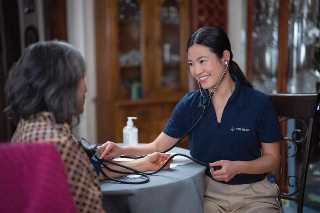 A VNS Health nurse taking her patient's blood pressure at a kitchen table.