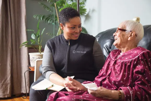 A VNS Health social worker smiling at and holding the hand of laughing senior woman.