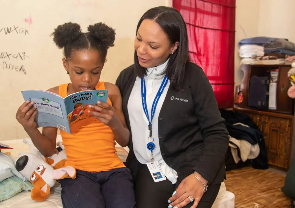 A VNS Health team member and a child sit on the corner of the bed while the child reads from a children's book.