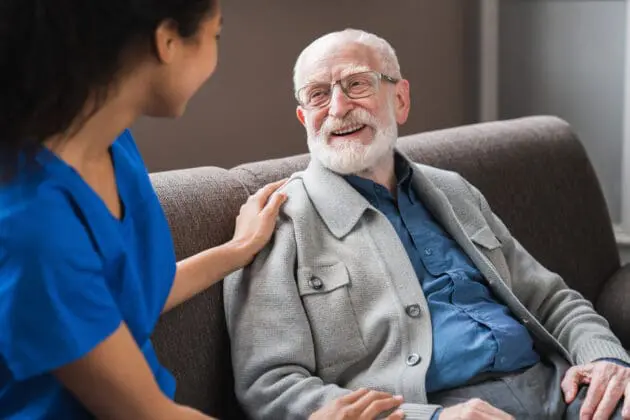 A nurse speaking with her elderly patient, gently resting her arm on his shoulder.