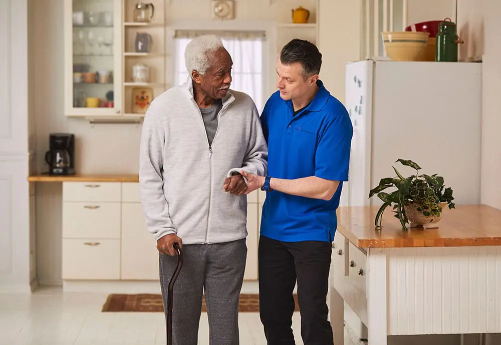 A VNS Health home health aide helping an elderly patient walk around his home.