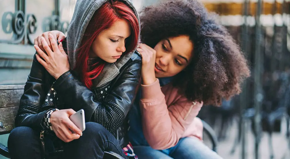 Teen with red hair holding a phone, comforted by a peer in a pink jacket, representing emotional support being offered through youth mental health services.