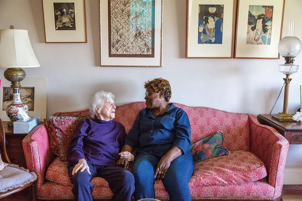 A VNS Health home health aide sitting with her patient on a red sofa.