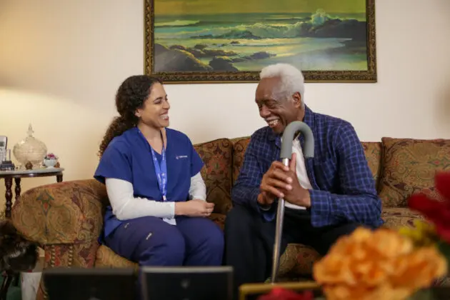 A VNS Health team member sitting on a couch with an elderly man smiling.