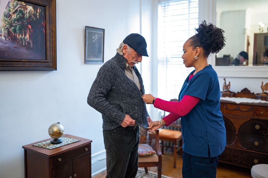 A senior man gets help zipping up his sweater by his home health aide.