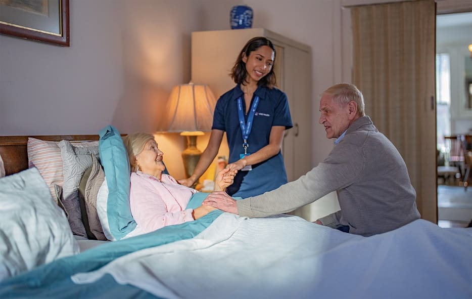 Senior woman smiling in bed with her husband holding one hand while a team member holds the other.