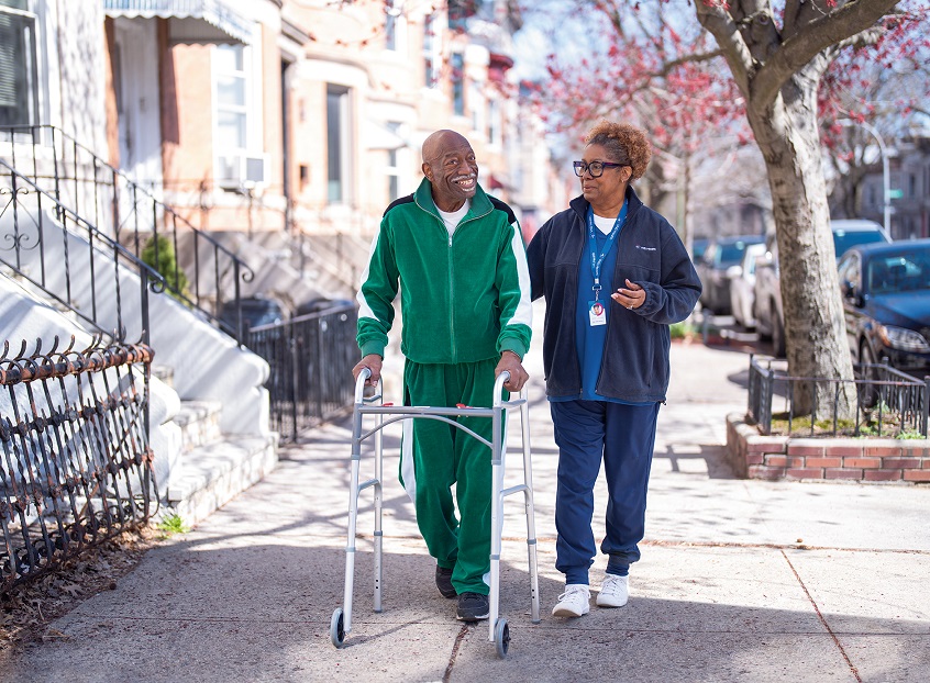 A home health aide walks outside with a senior man who needs the support of a walker.