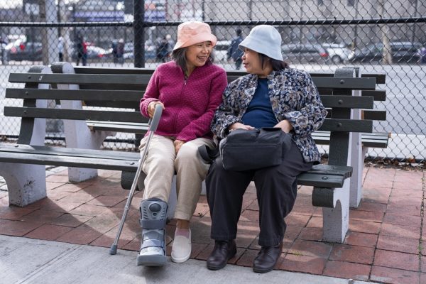 Two senior women sit on a bench. One woman has her foot in a boot and holds a cane.
