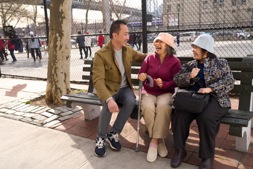 Picture of a senior Asian woman with a cane laughs as she sits between another senior Asian woman and a younger Asian man.