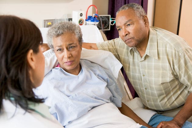 A concerned woman in a hospital bed and her husband listen to a doctor.