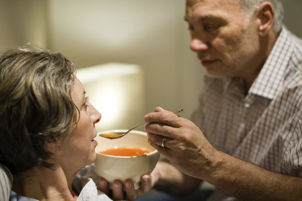 A husband feeds his wife tomato soup while she is propped up in bed.