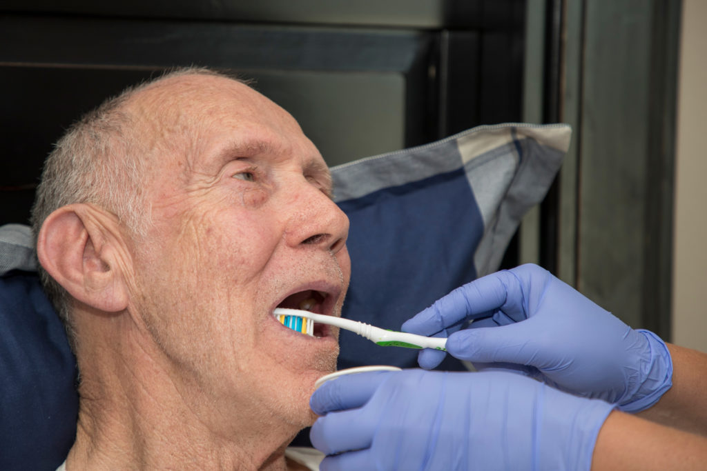 A senior man gets his teeth brushed by someone wearing latex gloves.