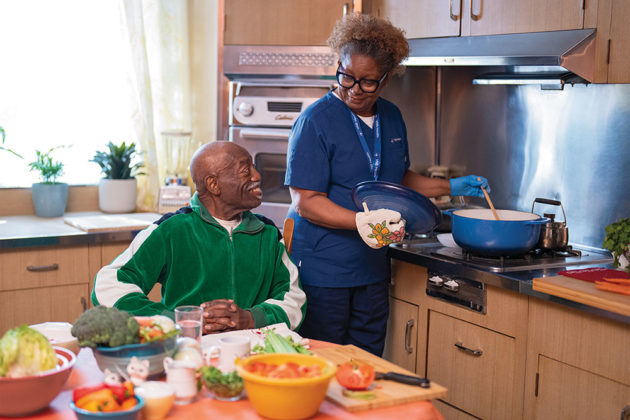 A home health aide stirs a pot in the kitchen for an older gentleman sitting at the table.