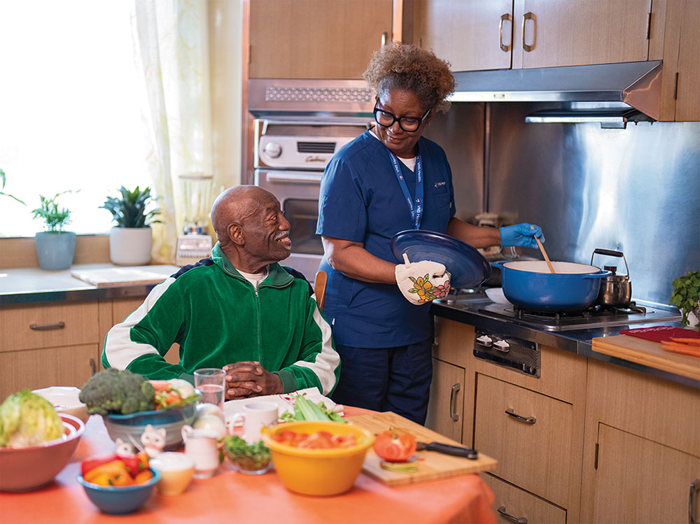 A home health aide stirs a pot in the kitchen for an older gentleman sitting at the table.