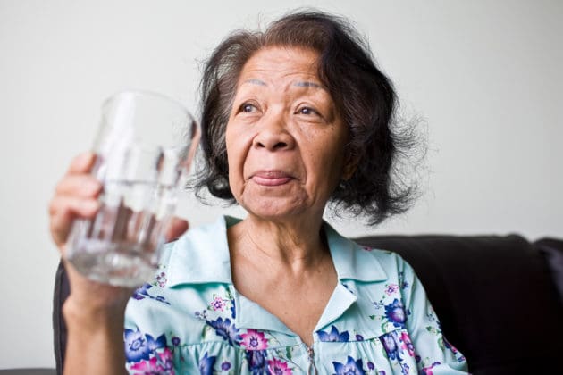 A senior Asian woman drinks a large glass of water.