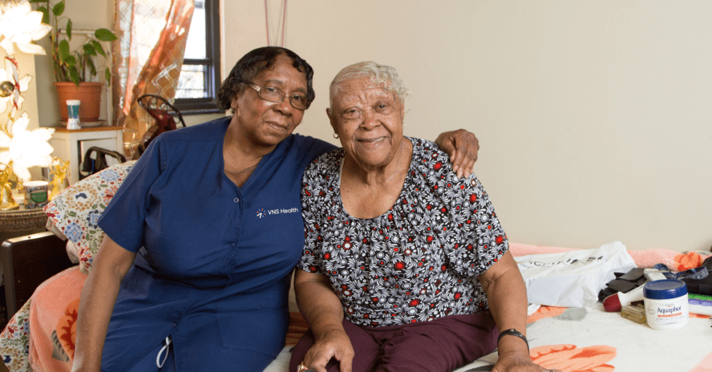 A VNS Health team member sits on a bed with her arm around an elderly woman.